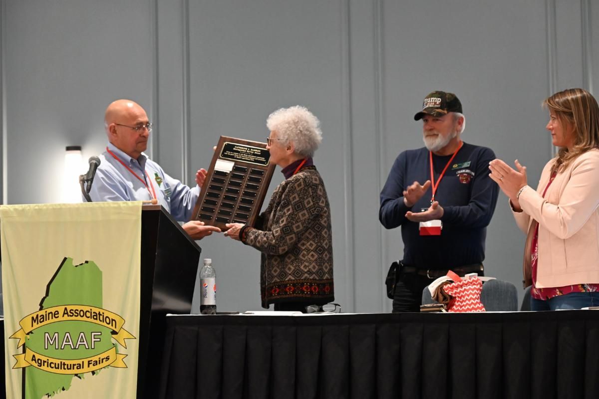 A presenter hands a plaque to a recipient at a Maine Association of Agricultural Fairs event, while others applaud.