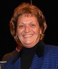 A smiling individual wearing a black turtleneck under a blue and black patterned blazer against a dark background.