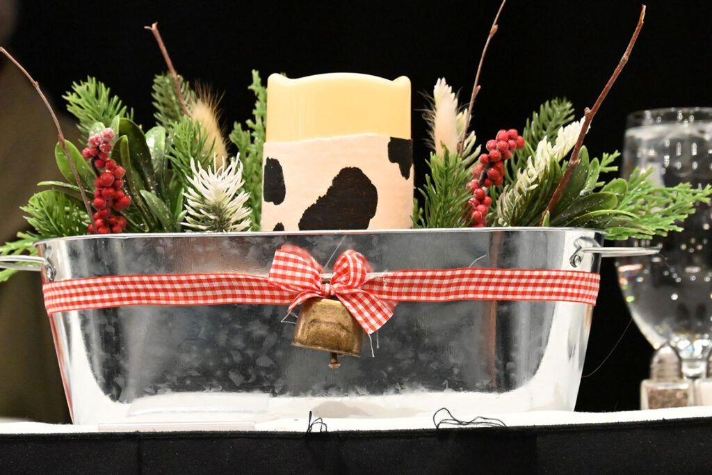 A galvanized metal trough centerpiece with evergreen, red berries, and a cow-print candle, tied with a red gingham ribbon.