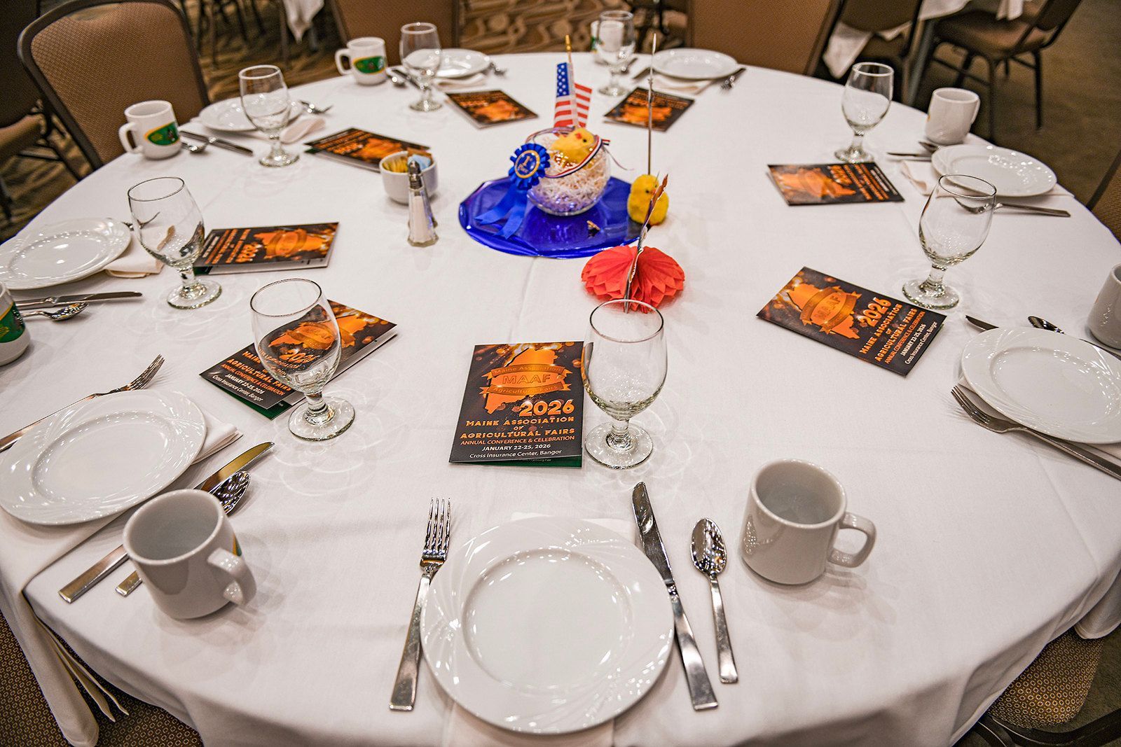 A square table set for a formal event with white linens, chairs in white covers, and yellow cloth napkins.