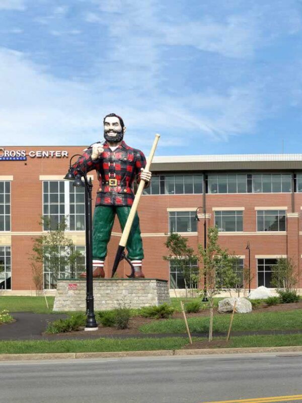 A large statue of a lumberjack in a red plaid shirt and green pants stands on a stone base in front of a brick building.