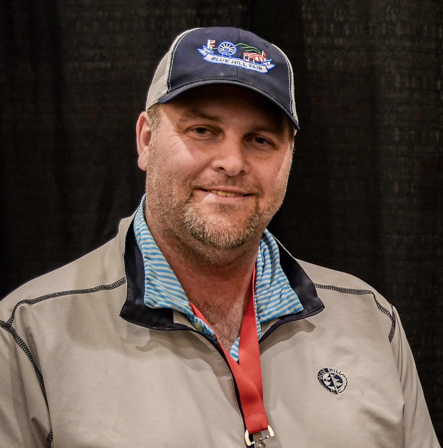 A person with light stubble wearing a dark baseball cap and a white quarter-zip jacket against a plain wall.