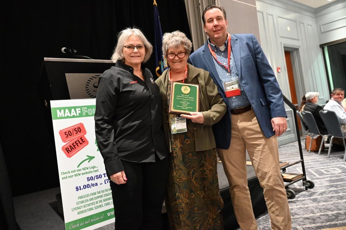 Three people stand on a stage next to a 50/50 raffle sign; one holds an award plaque while posing for a photo.