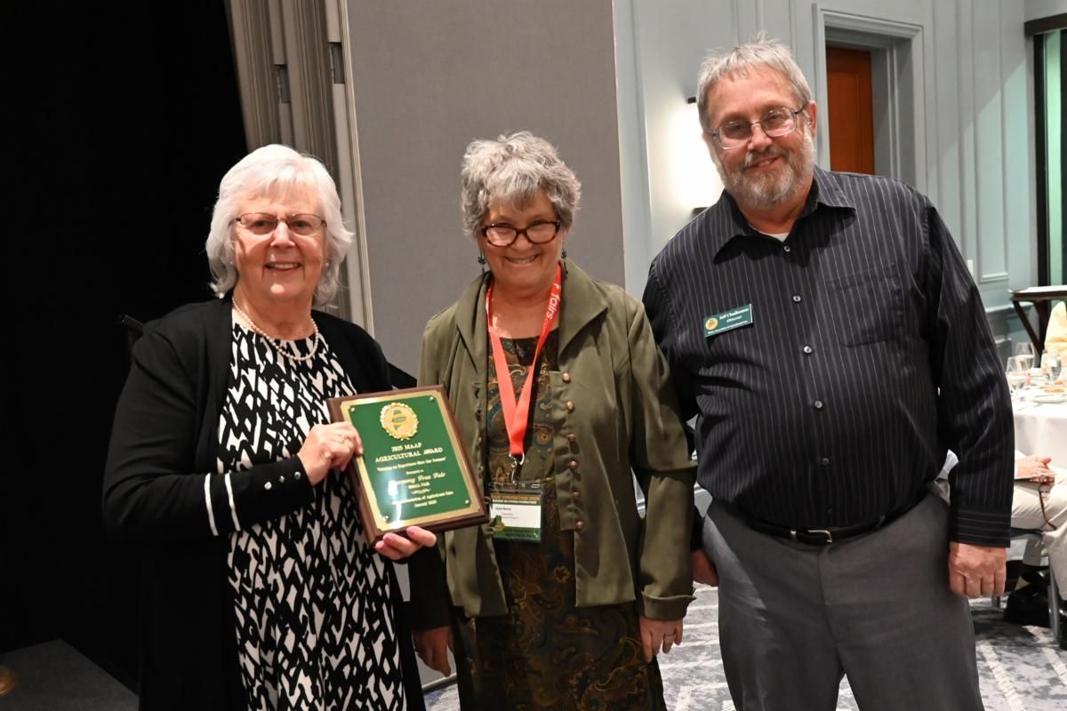 Three people stand together, with the person in the middle holding an award plaque at an indoor event.