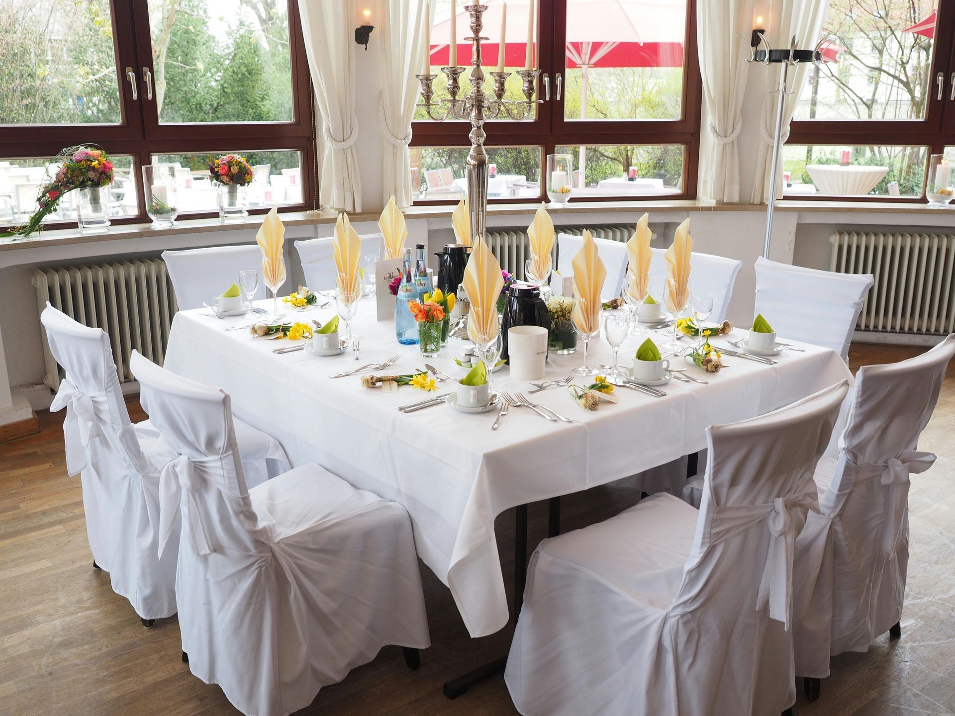 A square table set for a formal event with white linens, chairs in white covers, and yellow cloth napkins.