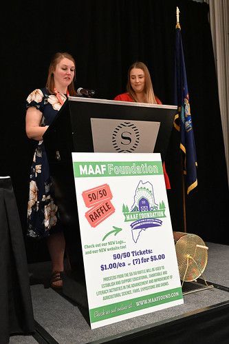 Two people stand at a podium on a stage with a Maine Aquaculture Association raffle sign, flag, and drum.