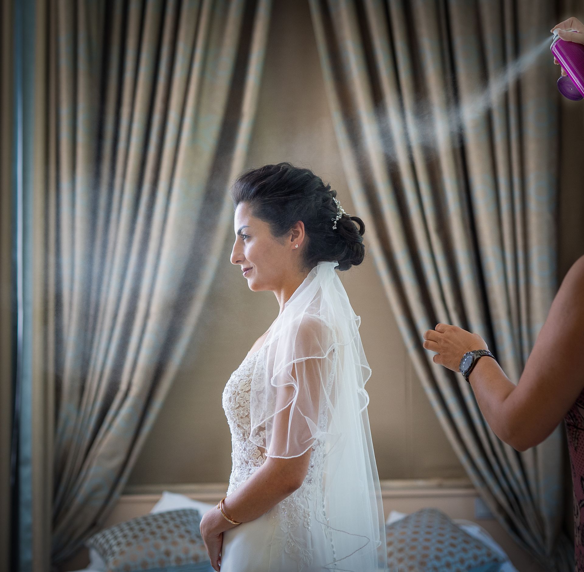 Braut bekommt Haarspray, im Brautkleid, in stilvollem Hotelraum – Getting Ready Hochzeitsfotografie NRW