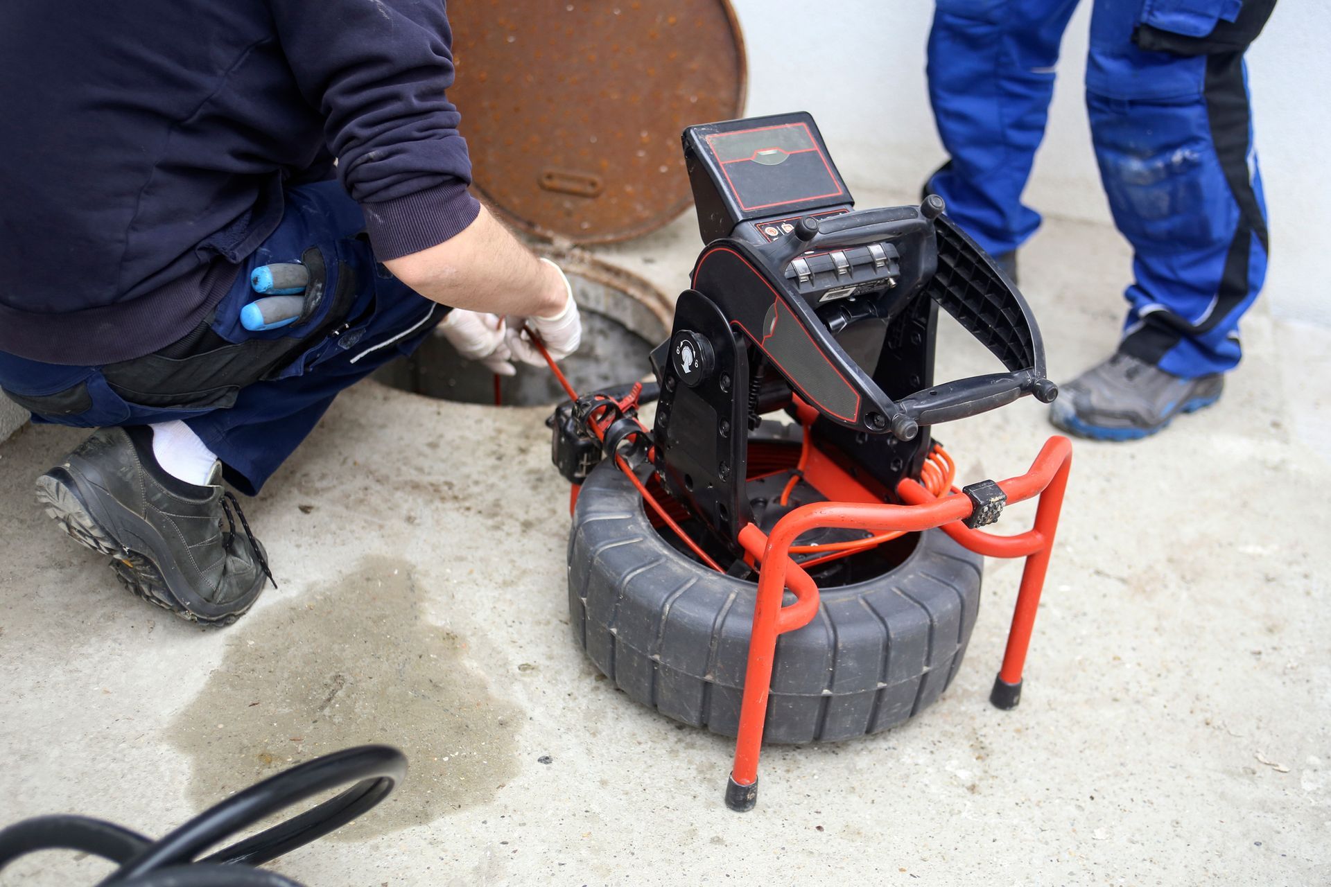 Workers using a sewer inspection camera to examine a manhole. The black and orange device sits on a tire, with a monitor visible.