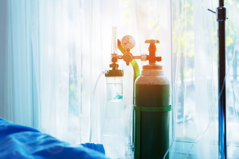 Hospital oxygen tank beside a bed in a bright room, with a window and IV stand nearby
