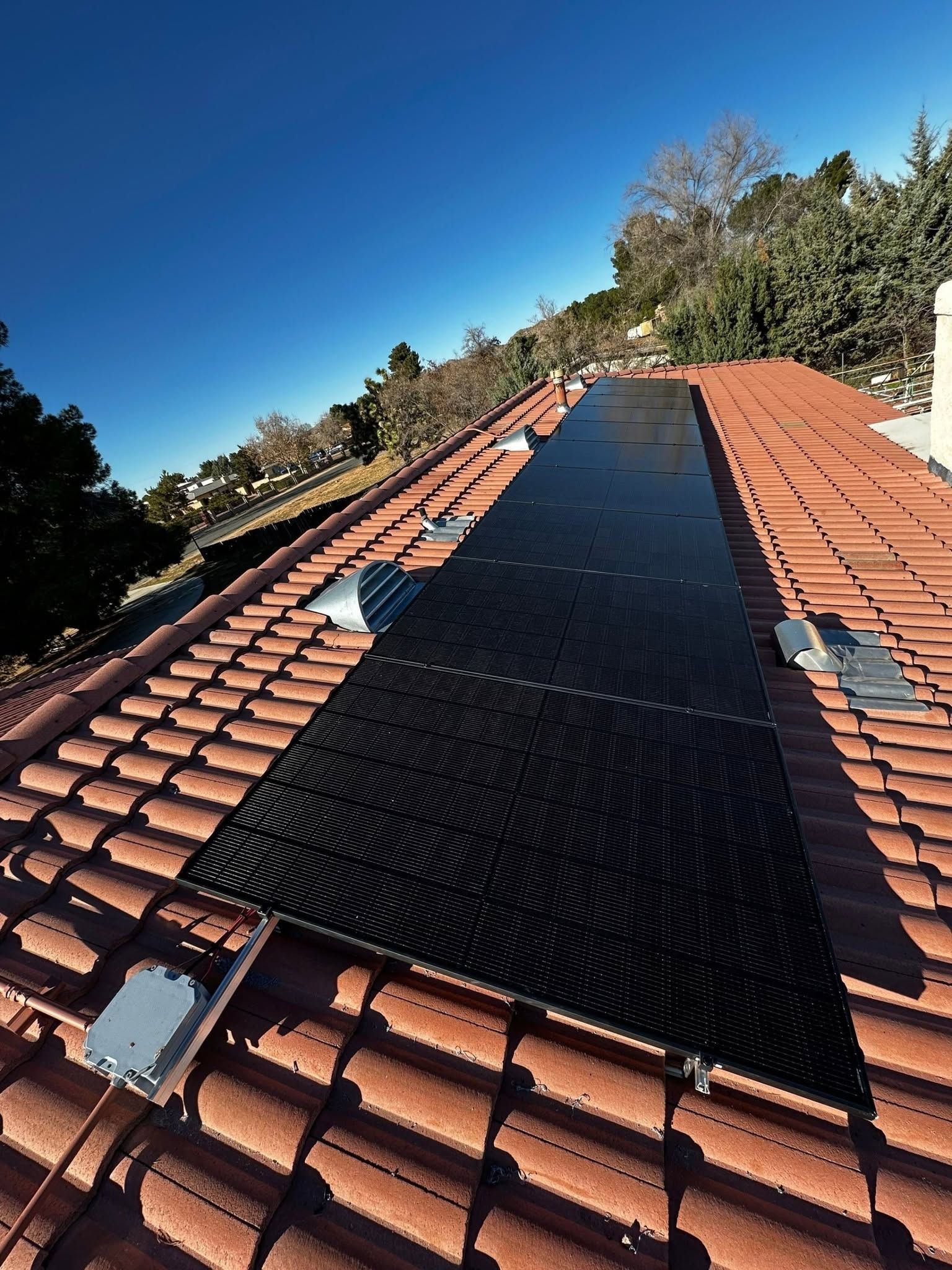 Roof with a large black solar panel array between red clay tiles under a clear blue sky.