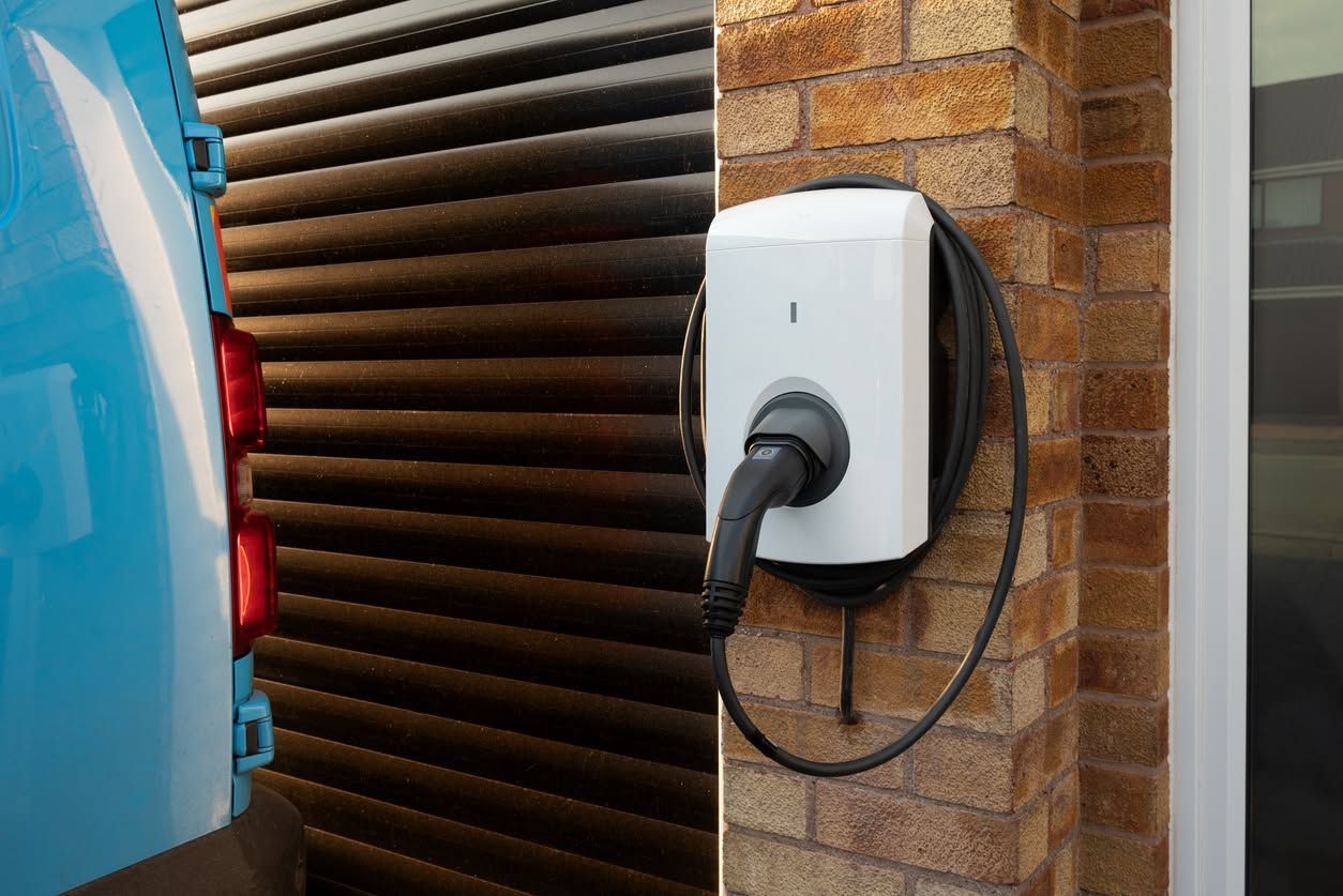 Electric vehicle charging port mounted on a brick wall beside a blue car.
