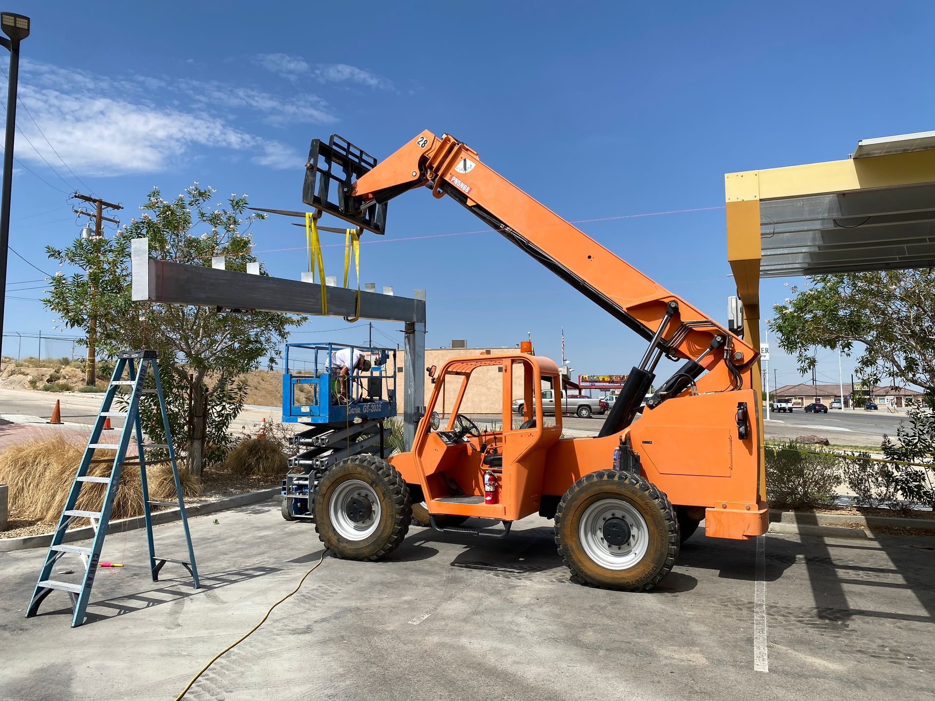 Orange telescopic forklift lifting a concrete beam beside a building under construction, with a ladder nearby.