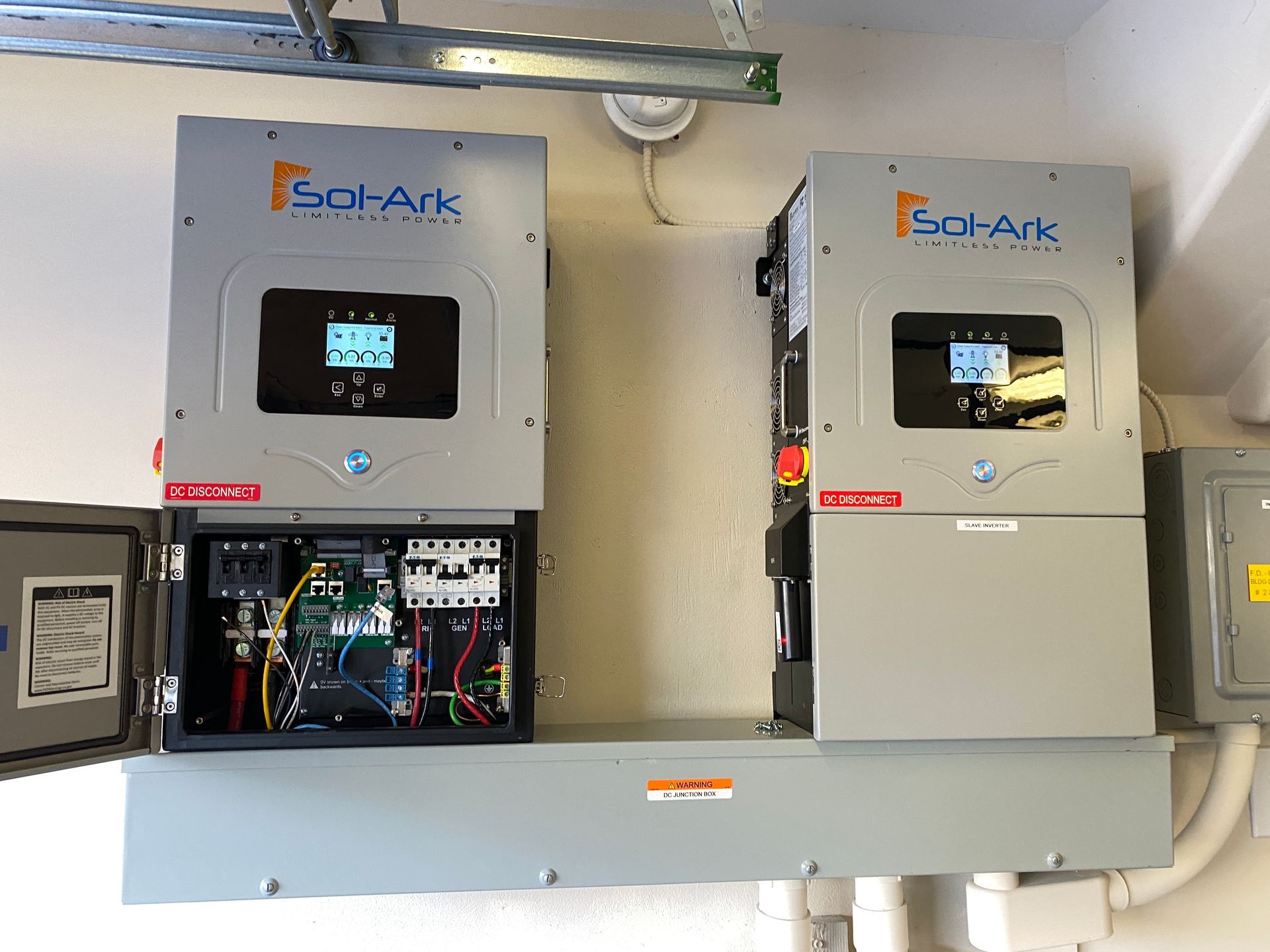 Two gray solar power inverters mounted on a wall with open electrical components below.