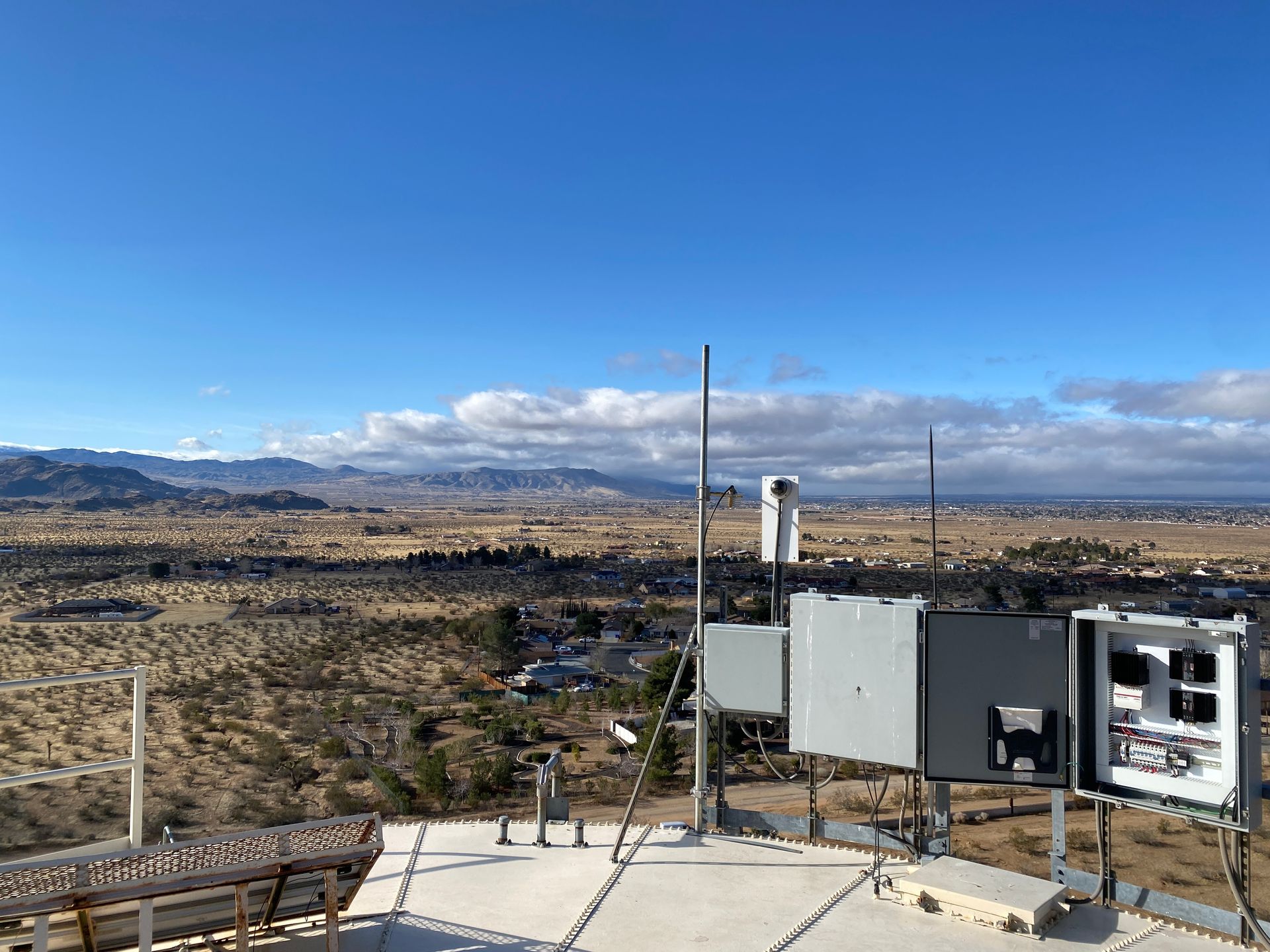Desert landscape with mountains under a blue sky, seen from a rooftop with equipment and railing.
