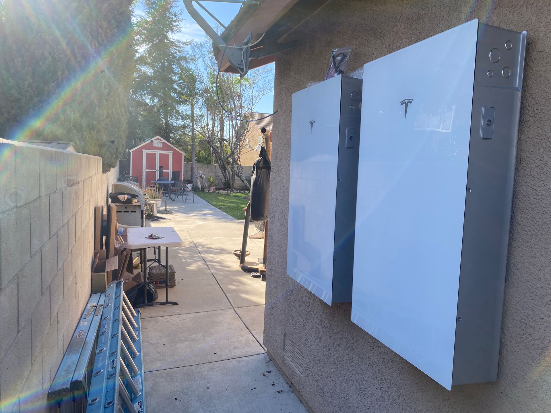 Narrow side yard with two white wall panels, a concrete path, tools, and a red shed in the distance.