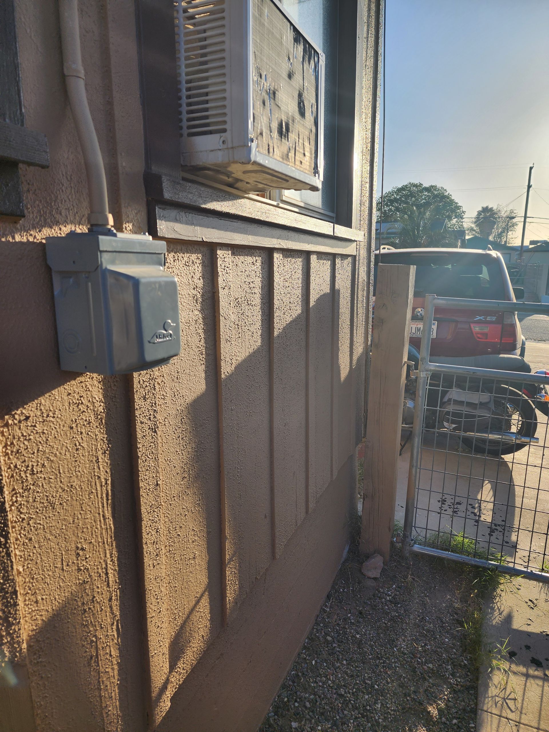 Side of a building with electrical boxes and a narrow walkway beside parked cars in sunlight