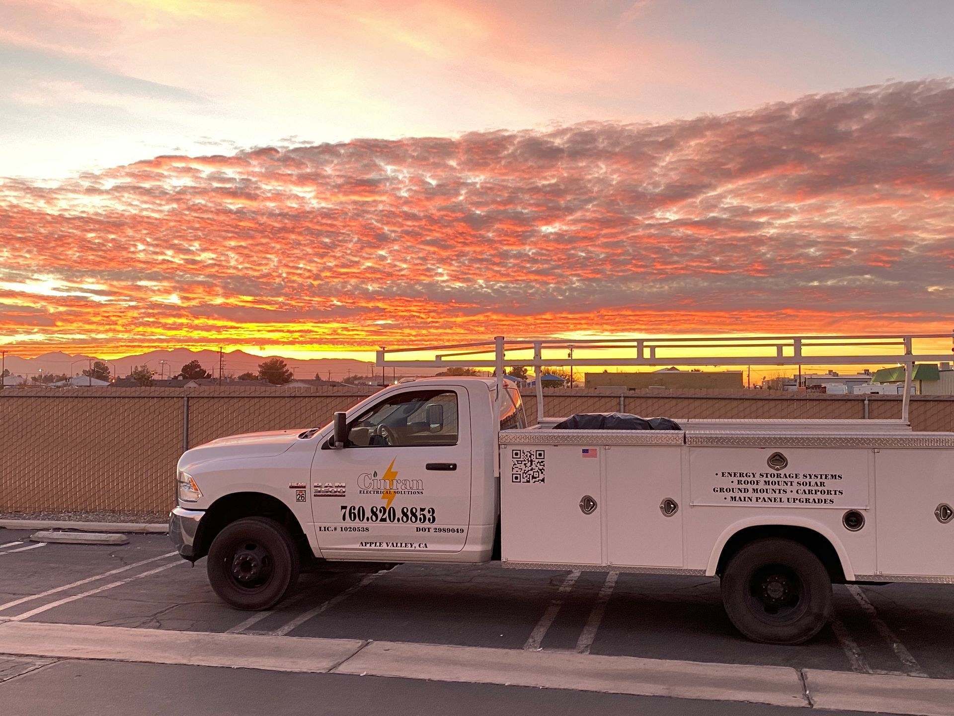 White utility truck parked by a fence under a colorful sunset sky.