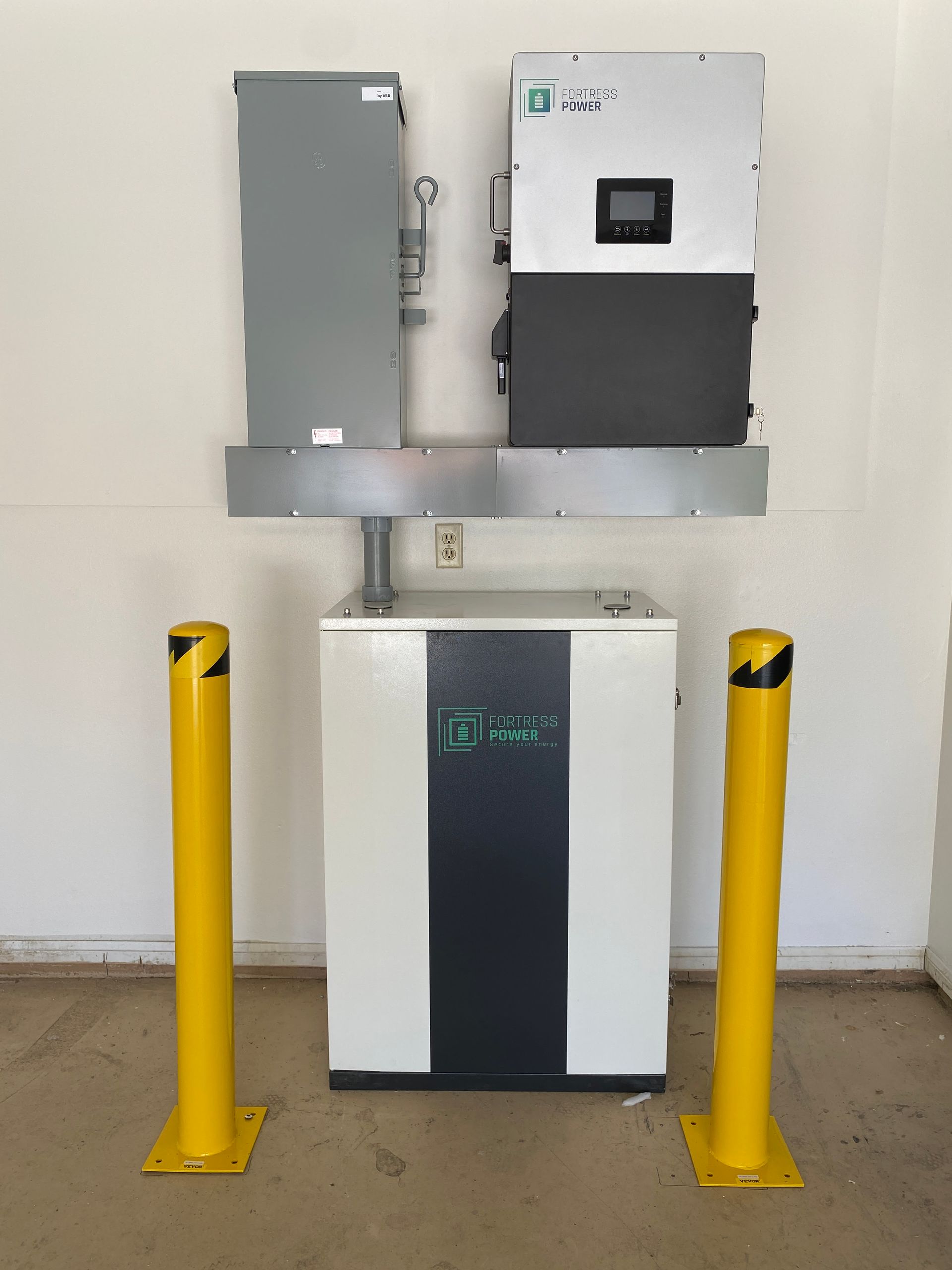 Wall-mounted electrical equipment with a white unit and gray cabinets, protected by yellow bollards.
