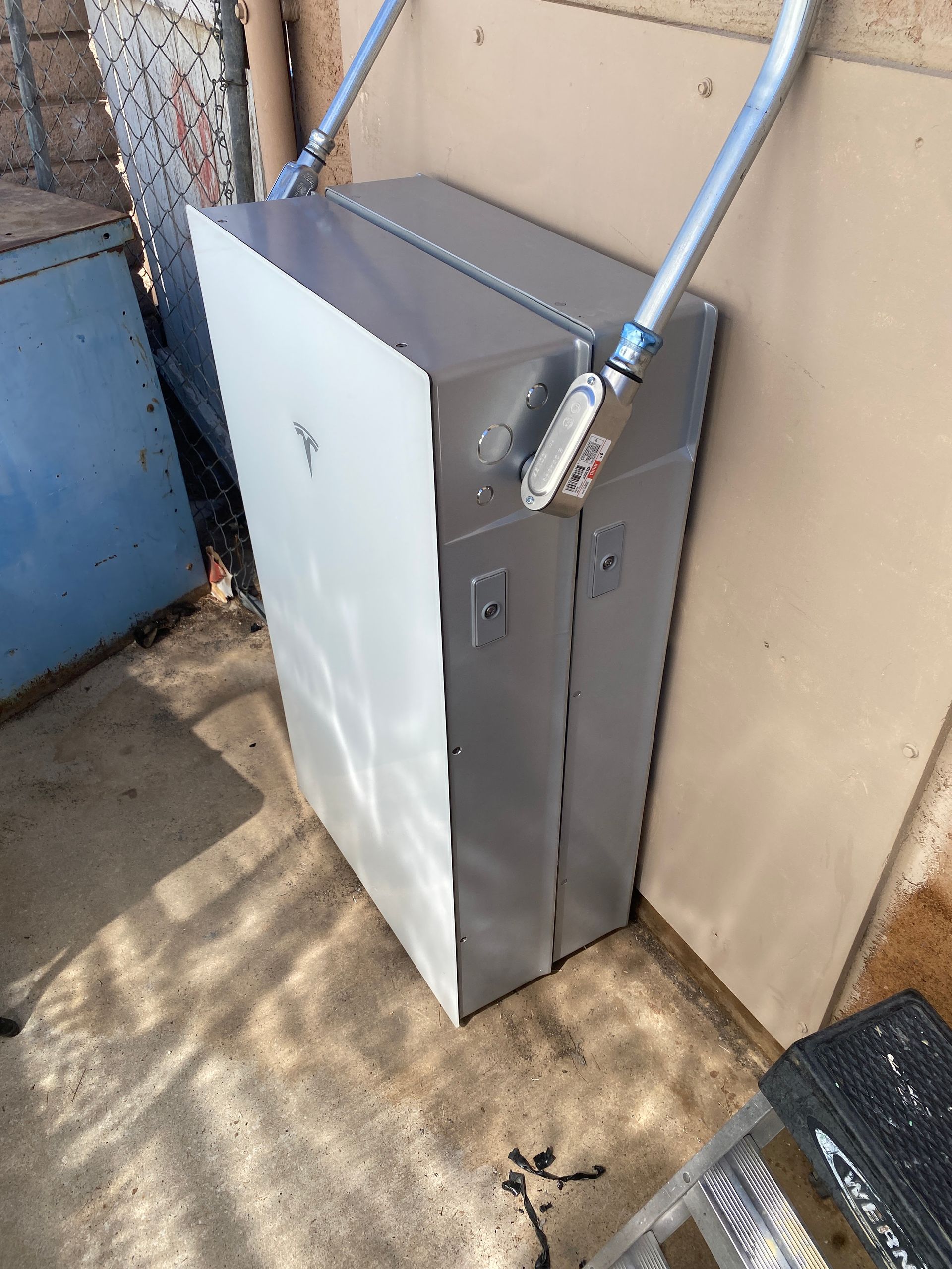 Gray electrical cabinet beside a wall with conduit pipes in an outdoor utility area.