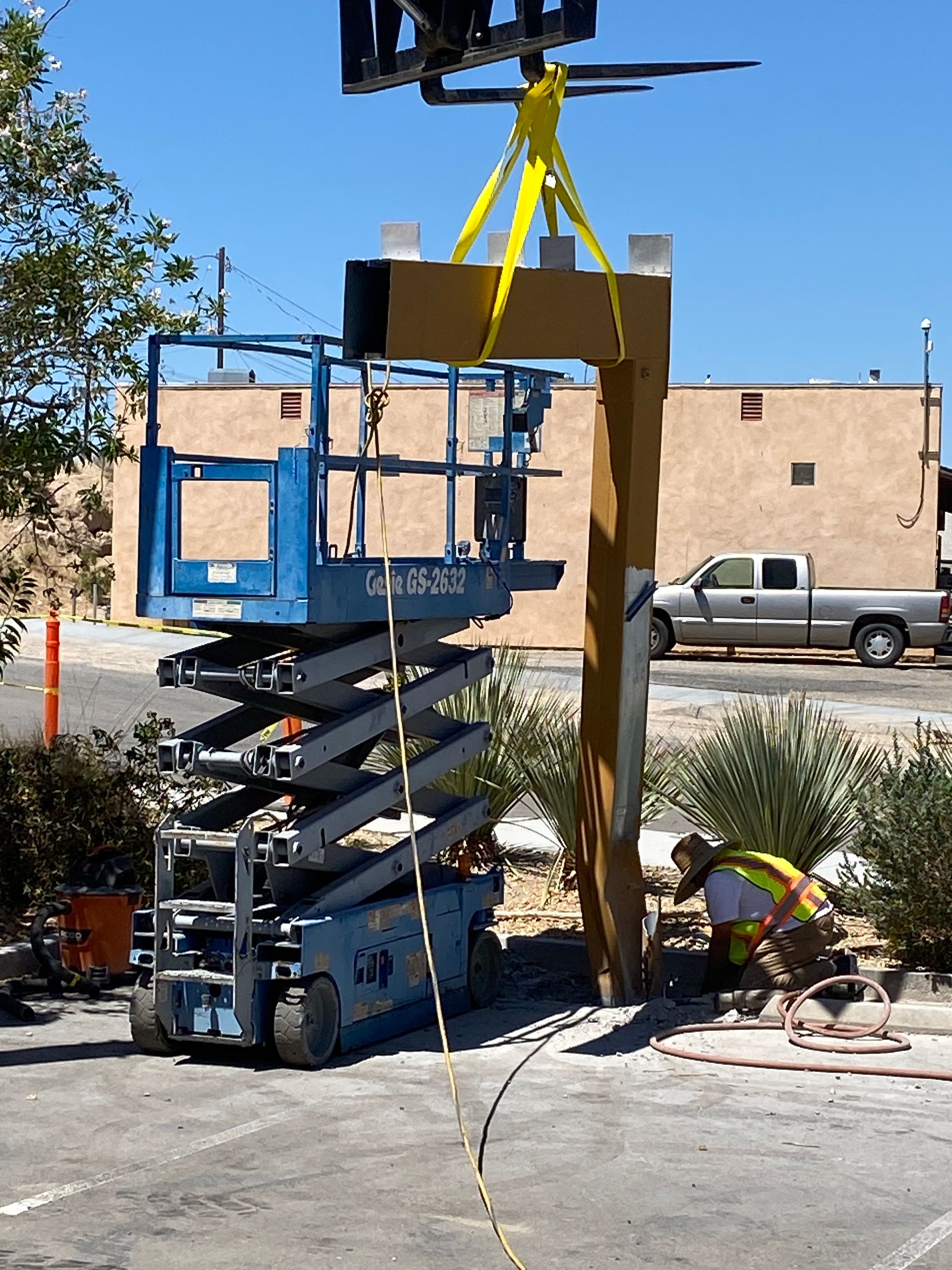 Blue scissor lift hoists a wooden frame with yellow straps beside a beige building.