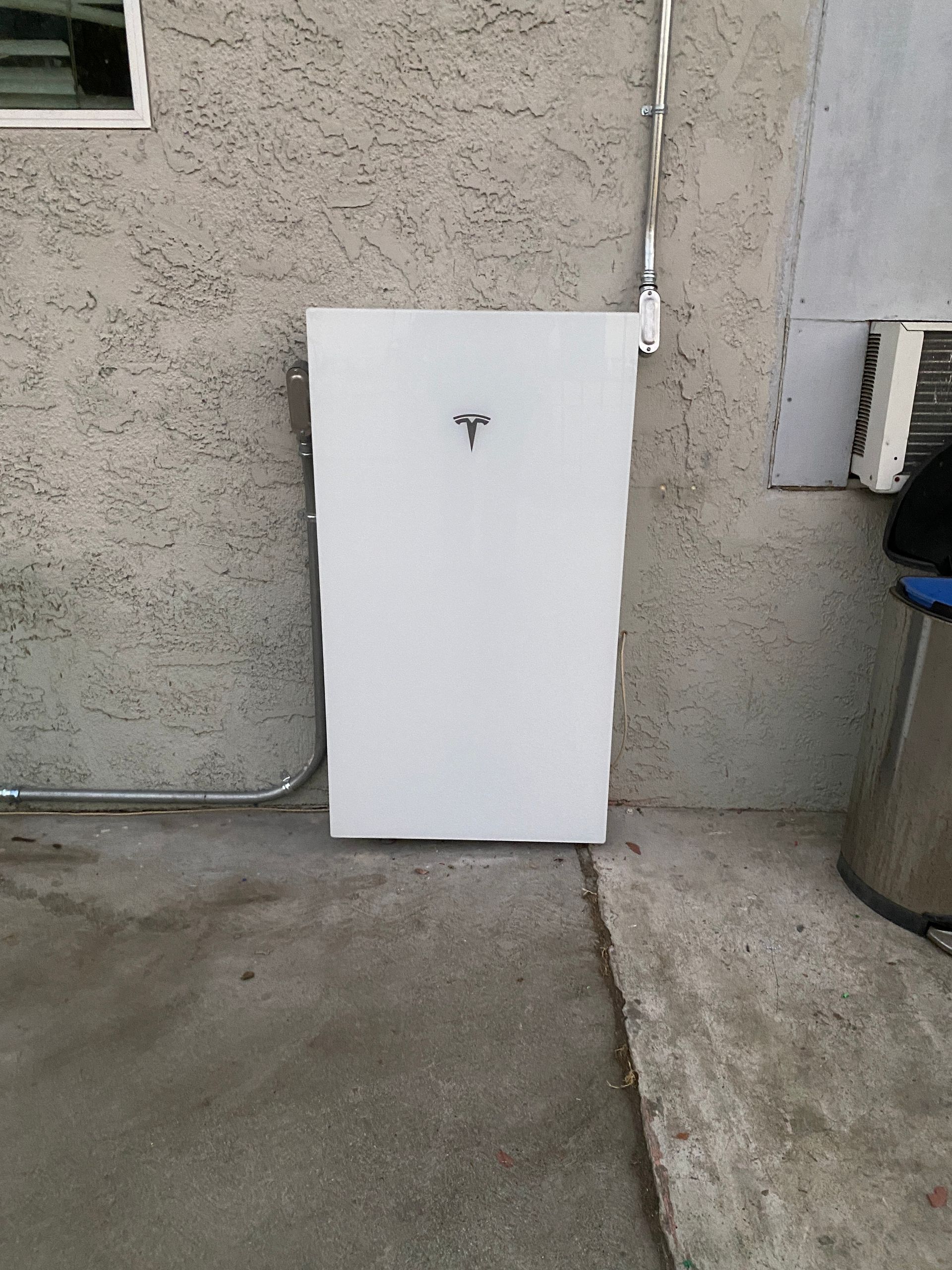 White utility panel leaning against an exterior stucco wall beside a concrete patio and trash can.