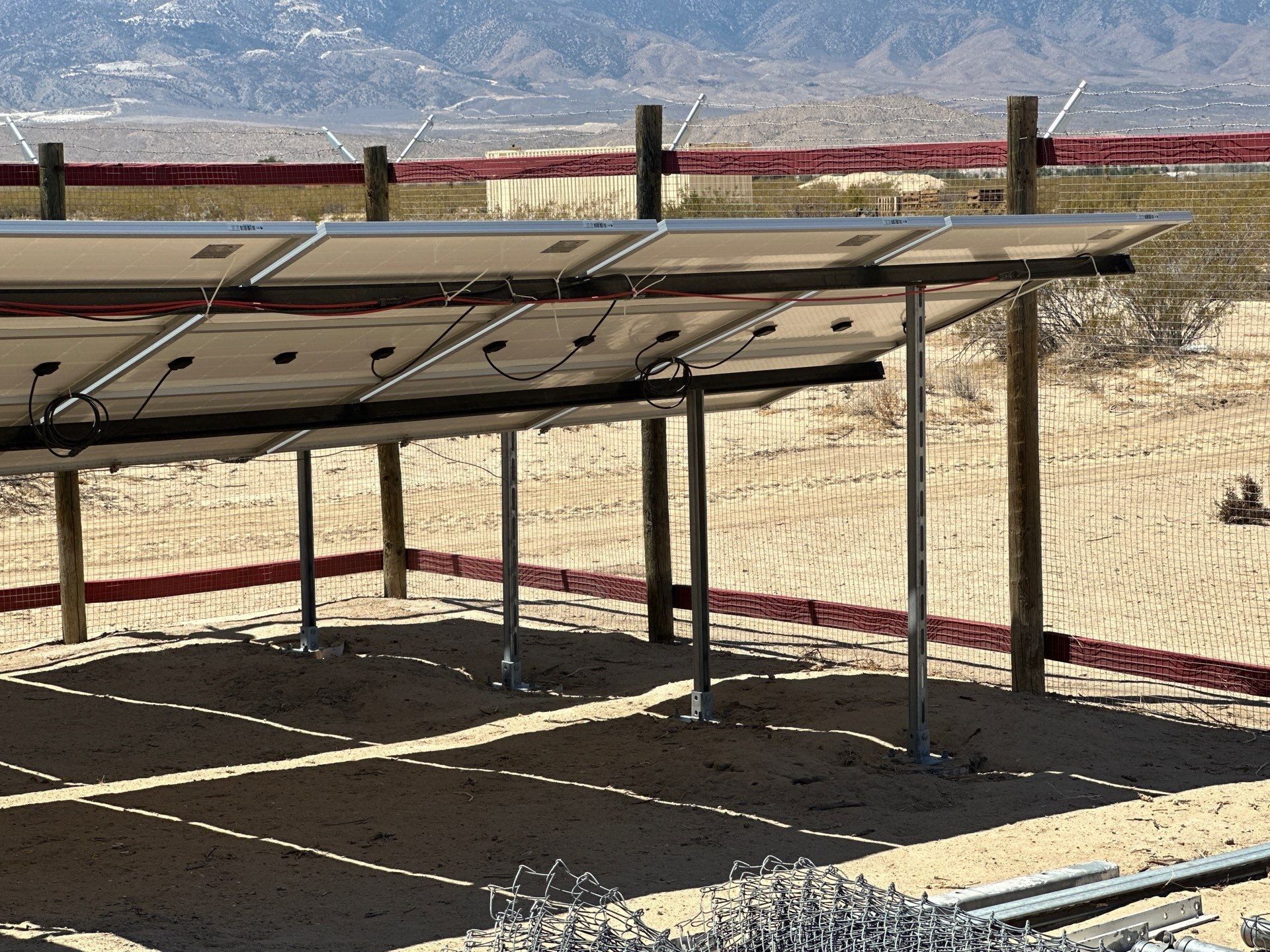 Solar panels mounted on a metal frame in a dry, sandy outdoor area with mountains in the background