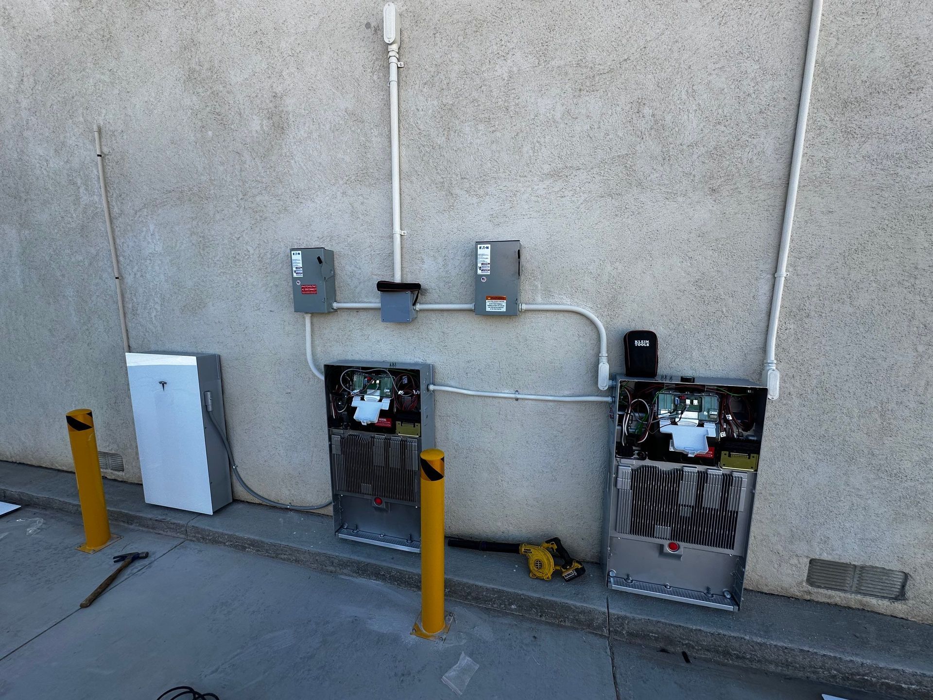 Outdoor utility meters and electrical boxes mounted on a stucco wall beside yellow bollards.