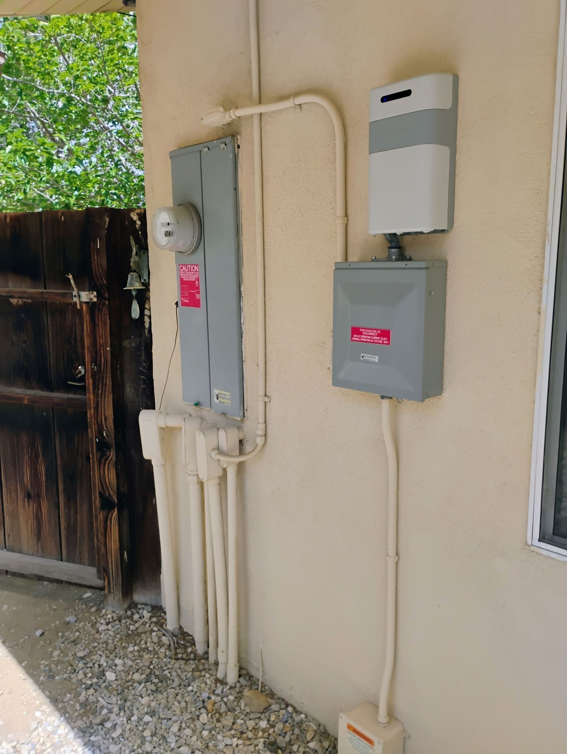 Utility meters and electrical boxes mounted on a beige exterior wall beside a fence.