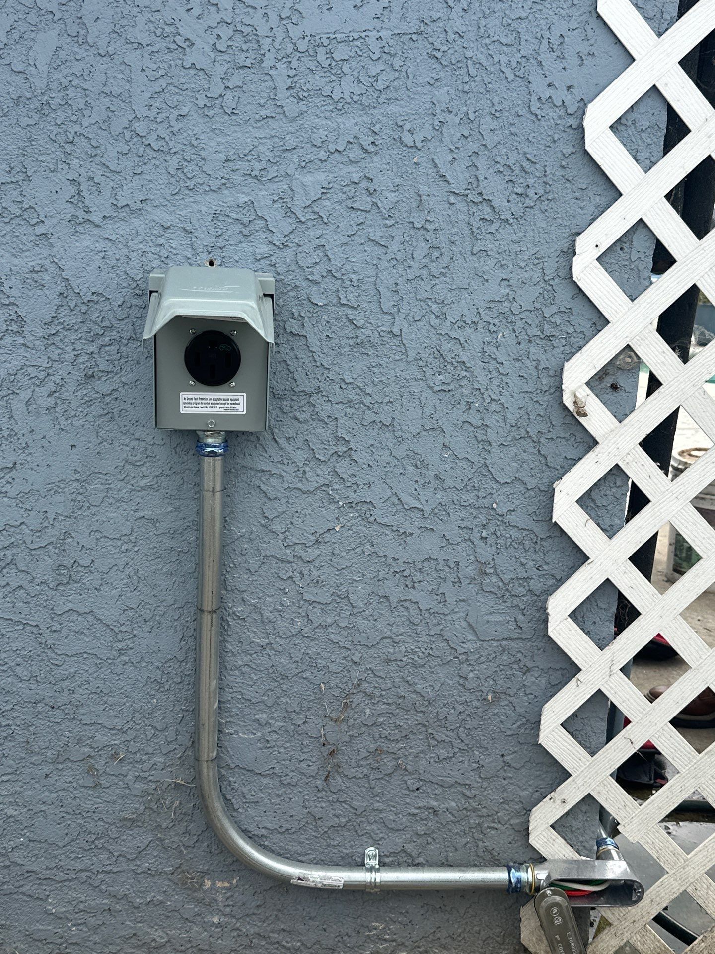 Gray electrical box on stucco wall with a metal conduit and white lattice panel nearby
