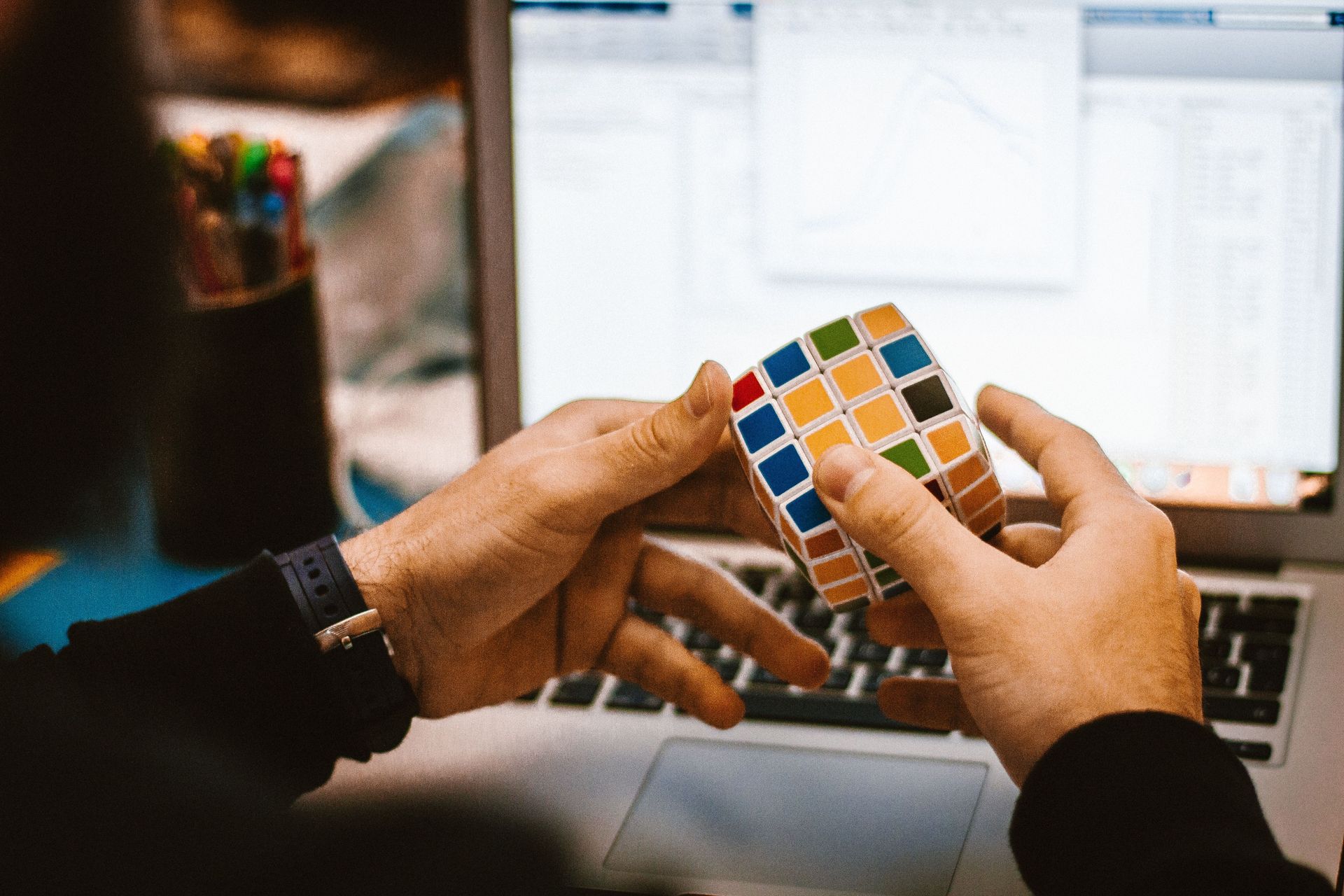 Hands holding a Rubik’s cube over a laptop keyboard, with a blurred computer screen in the background