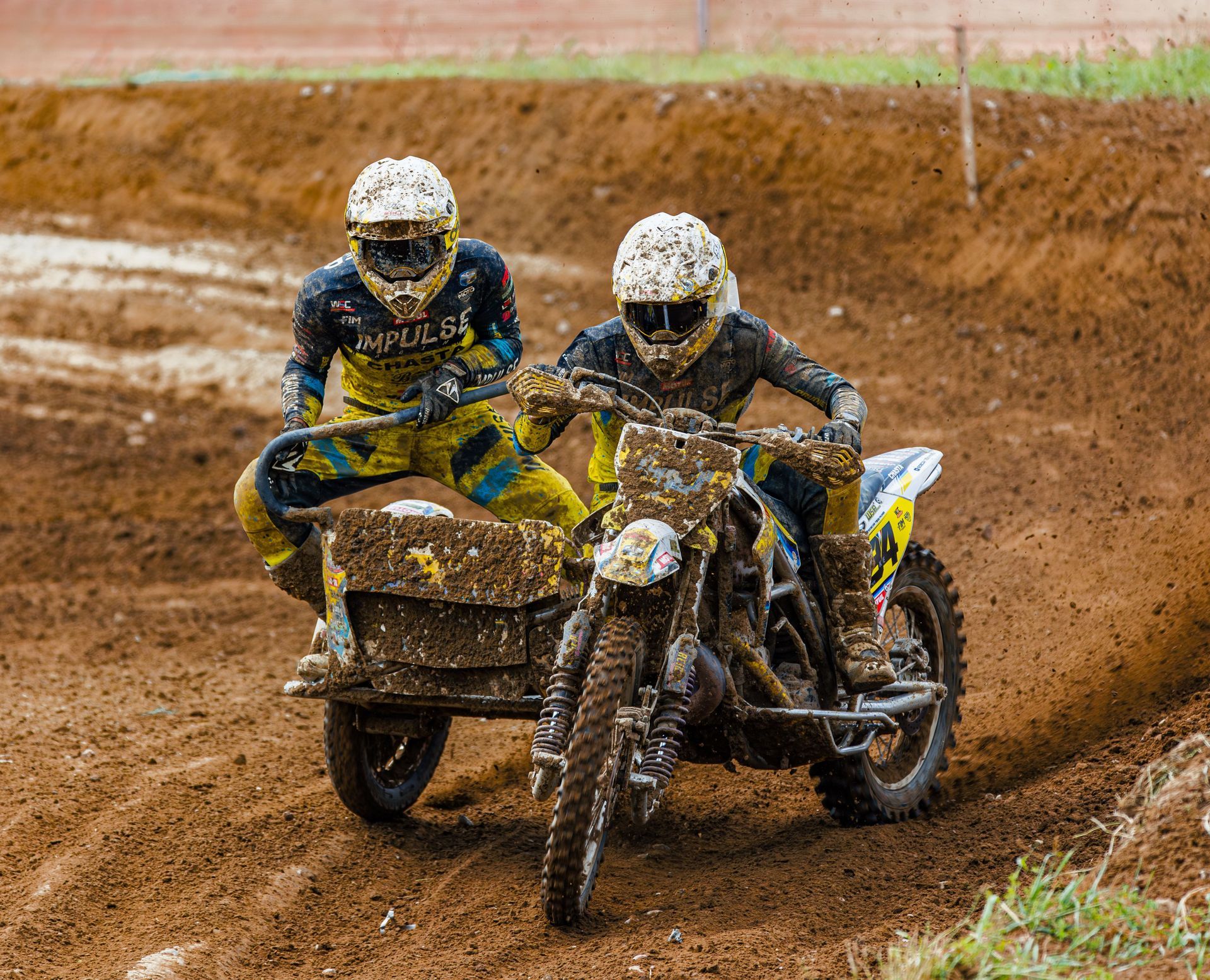 Two sidecar motocross riders race through a muddy dirt track, splashing mud.