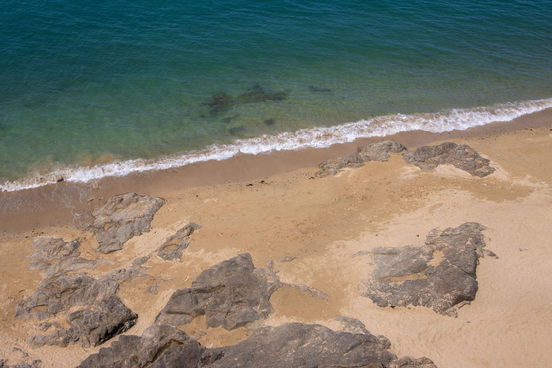 Aerial view of a sandy beach with clear turquoise water and scattered rocks.