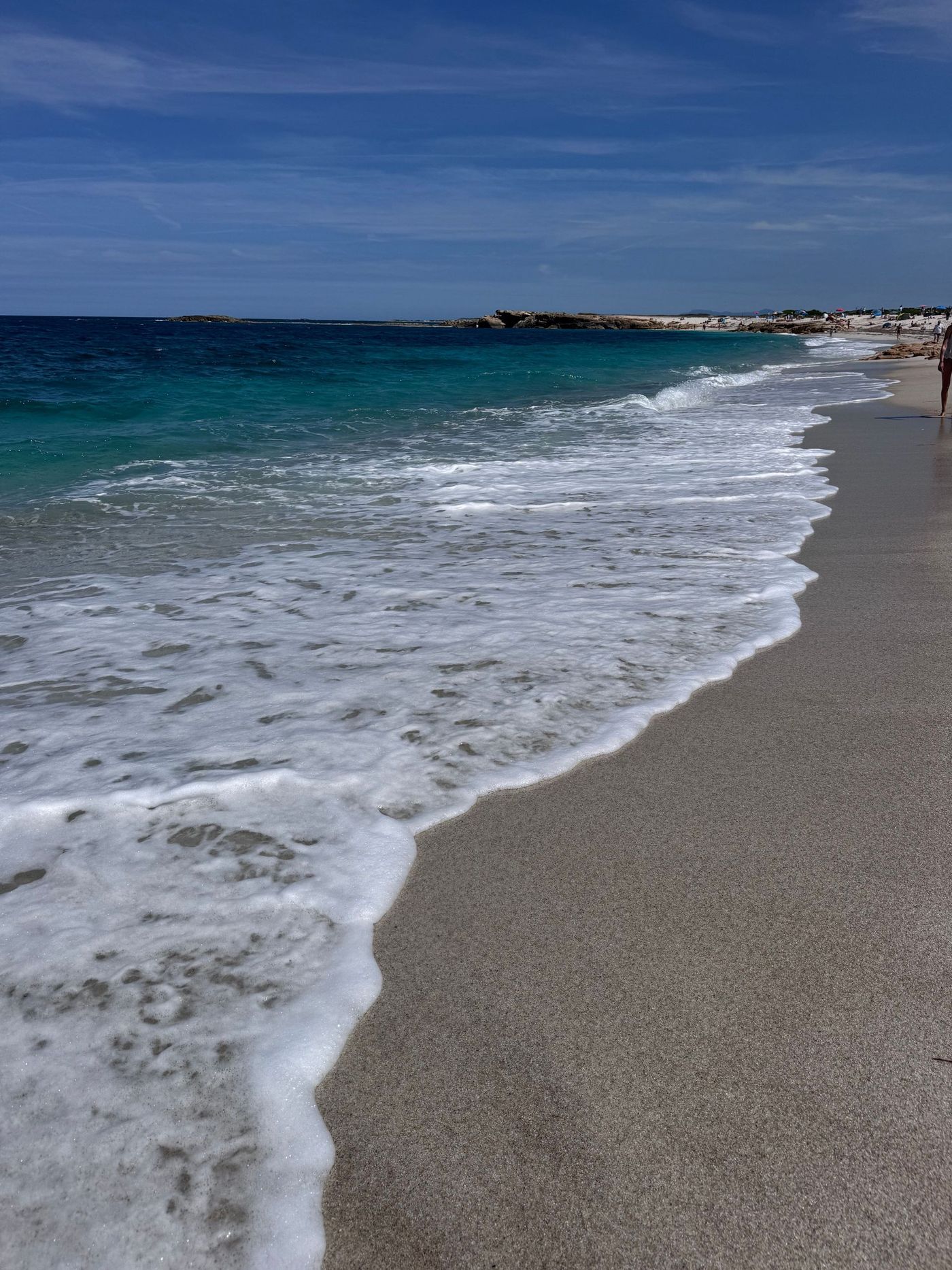 Waves washing onto a pebbled beach under a blue sky, with a distant shoreline in the background