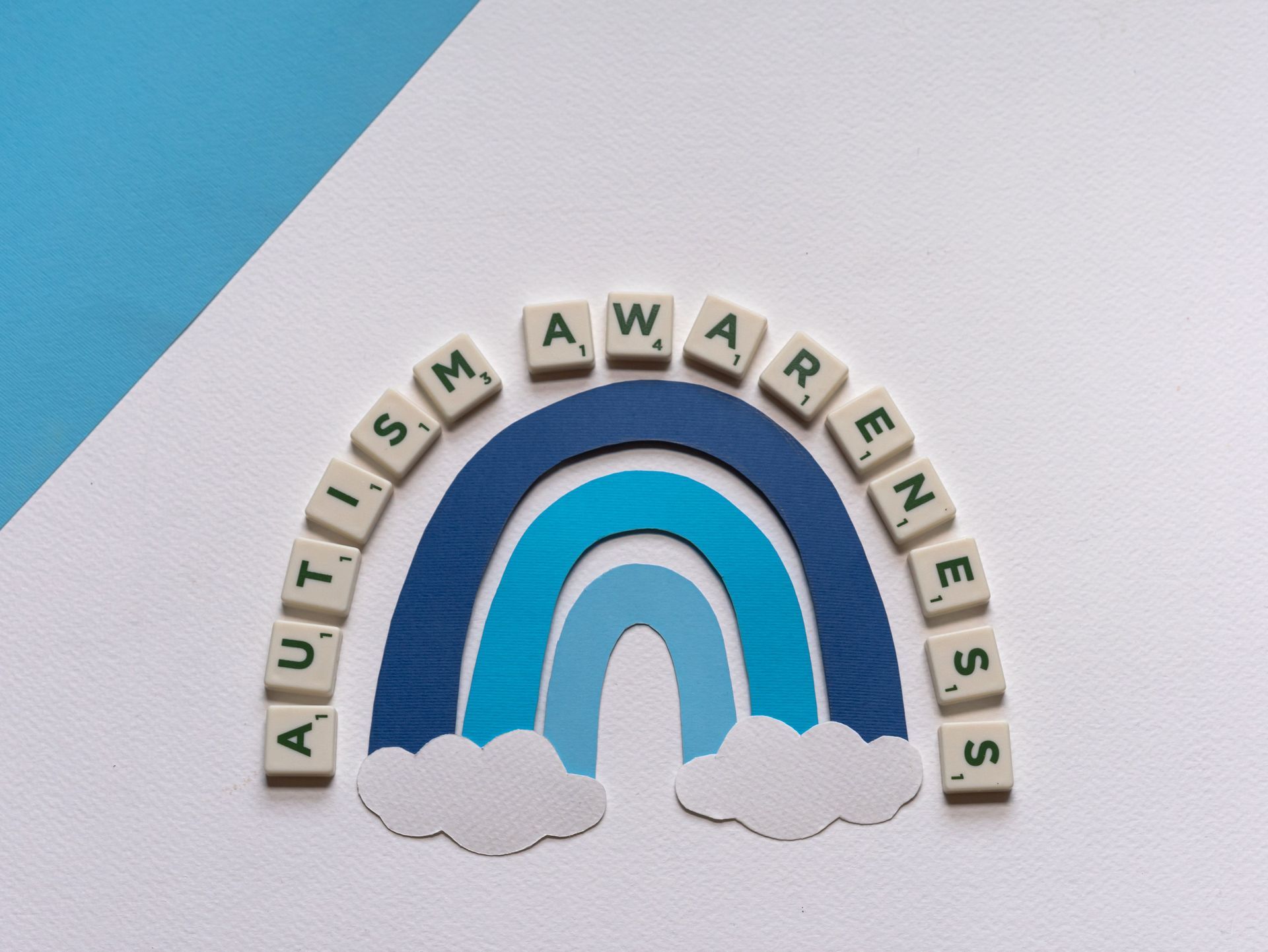 Rainbow with clouds and letter tiles spelling “autism awareness” on a blue and white background