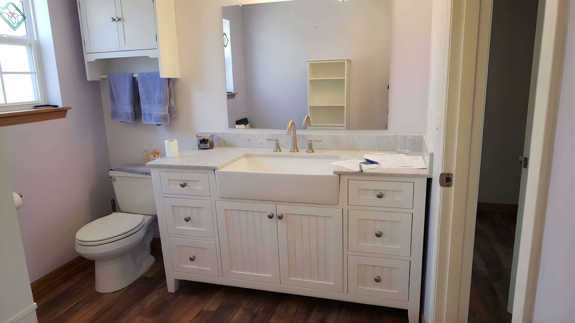 Bathroom vanity with white cabinets, sink, mirror, toilet, and a window with blue towels
