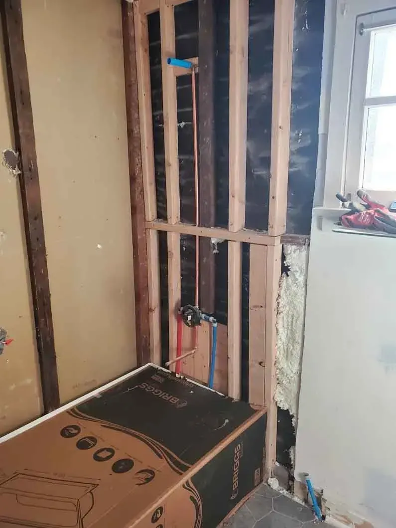 Partially gutted interior wall with exposed studs, wires, and a cardboard box near a window.