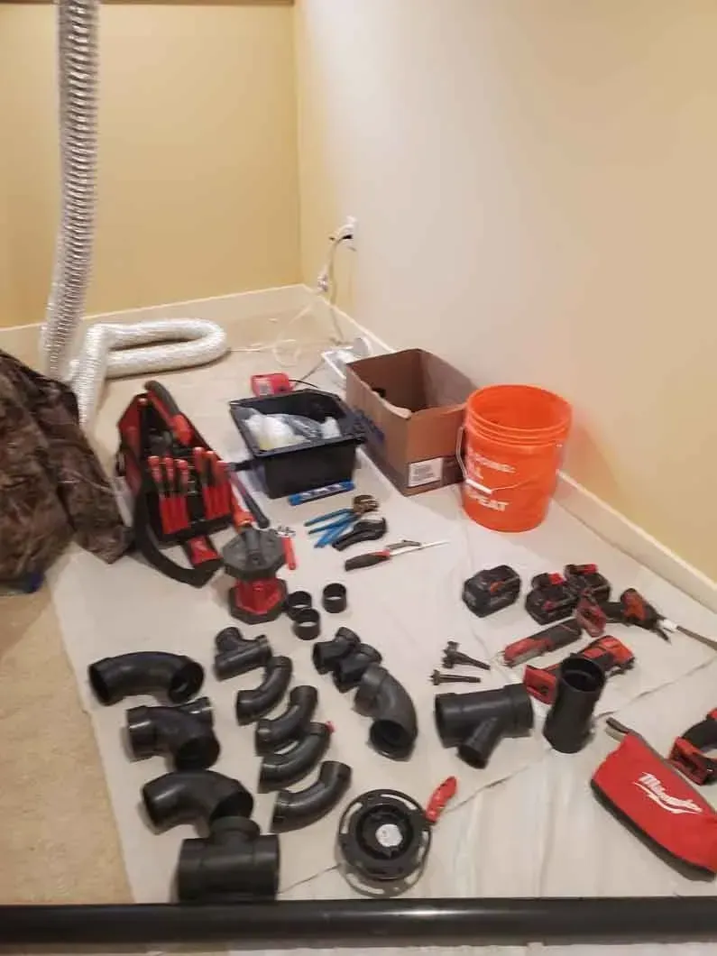 Assorted tools and hardware parts spread on a carpeted floor beside a wall and orange bucket.