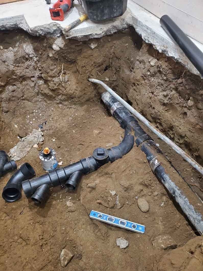 Excavated trench with black plumbing pipes and fittings laid in dirt beside a concrete edge.