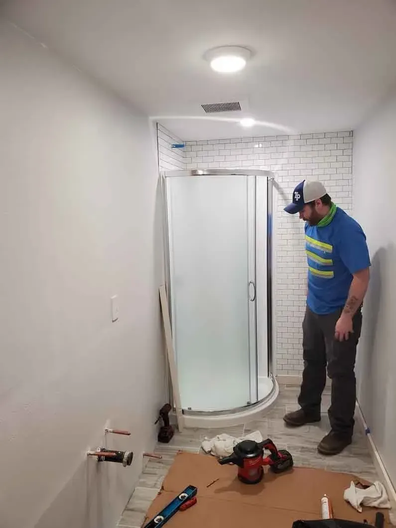 Worker in a small bathroom under renovation, standing beside a shower enclosure with tools on the floor.