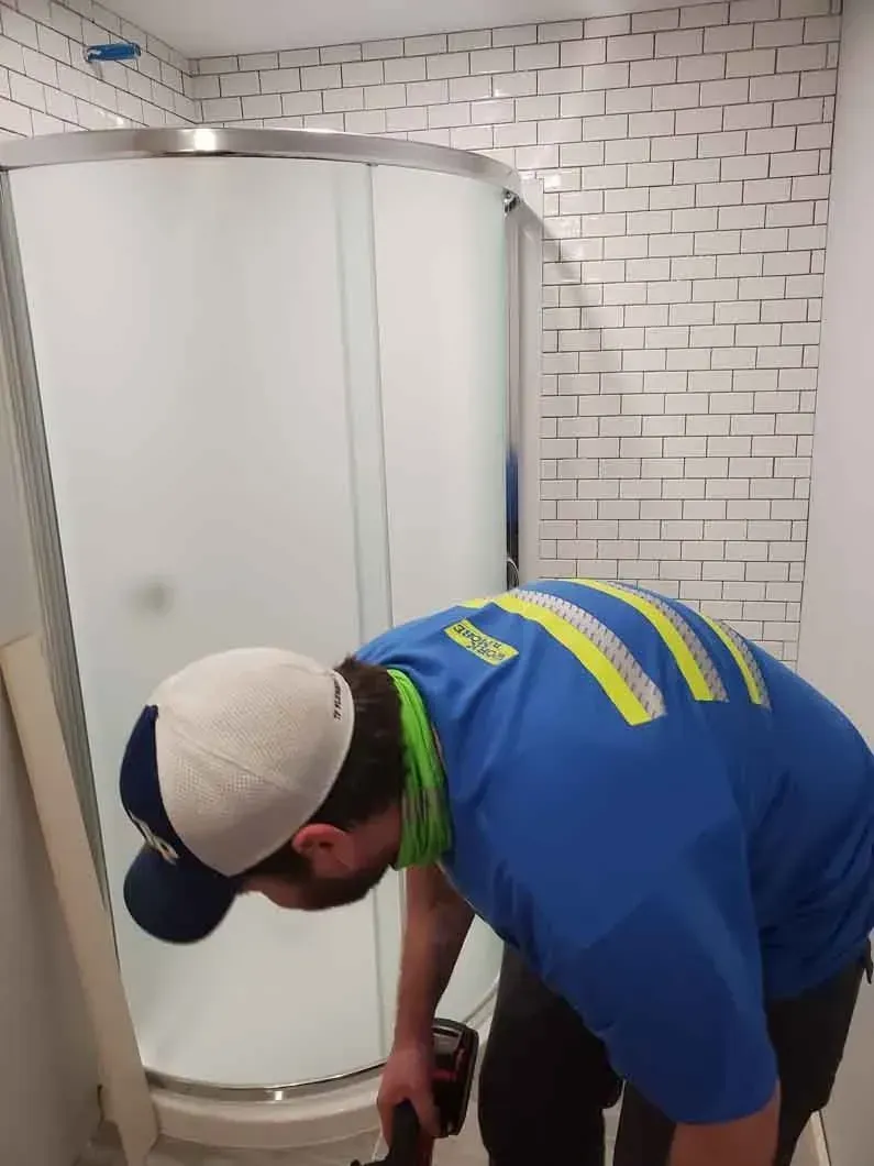 Worker in blue shirt bending over beside a curved shower enclosure in a tiled bathroom.