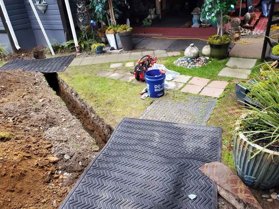 Garden path under construction with a trench, gravel, and a blue bucket near potted plants.