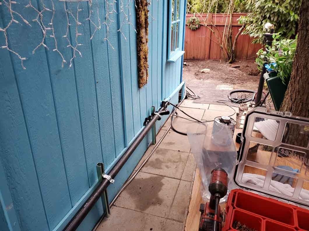 Narrow blue-painted walkway beside a house, with garden tools and a hose leading to a yard gate.