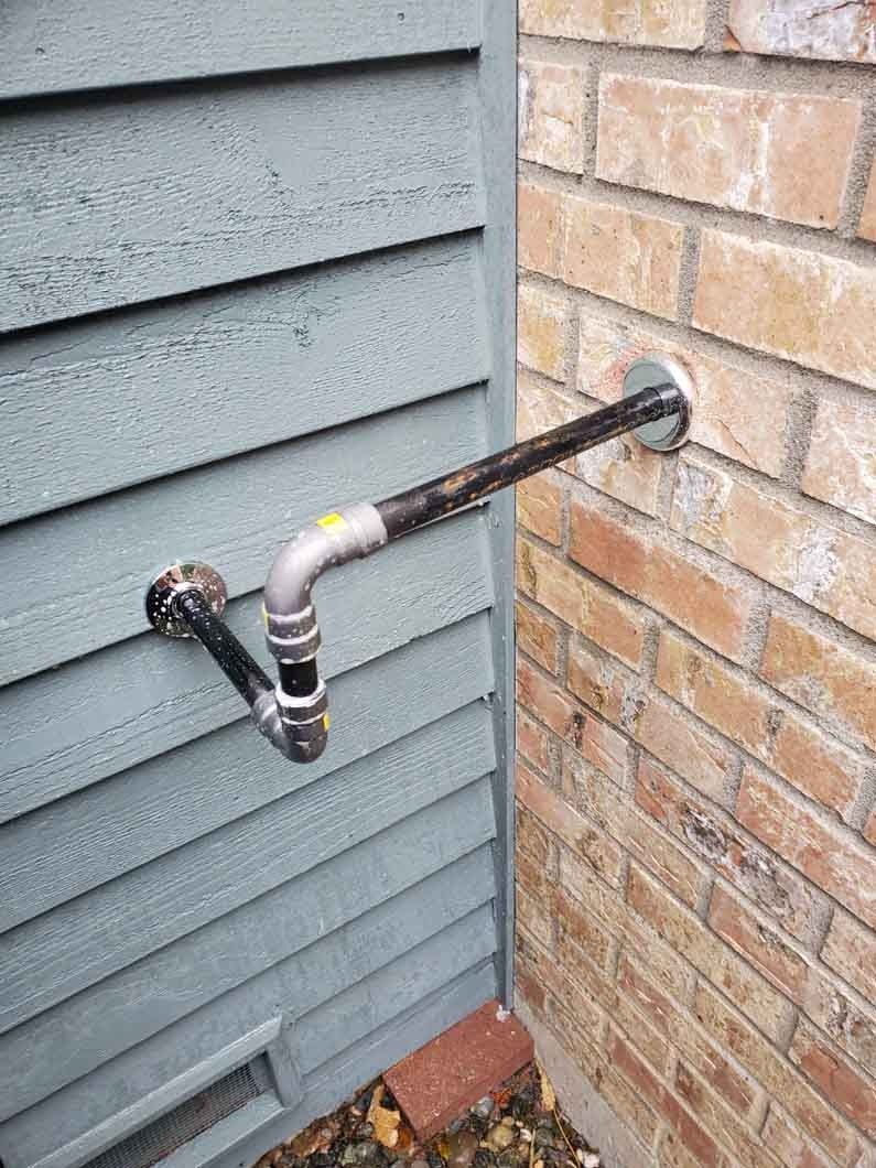 Plumbing pipe runs from brick wall to gray siding, with an elbow joint and valve outdoors.
