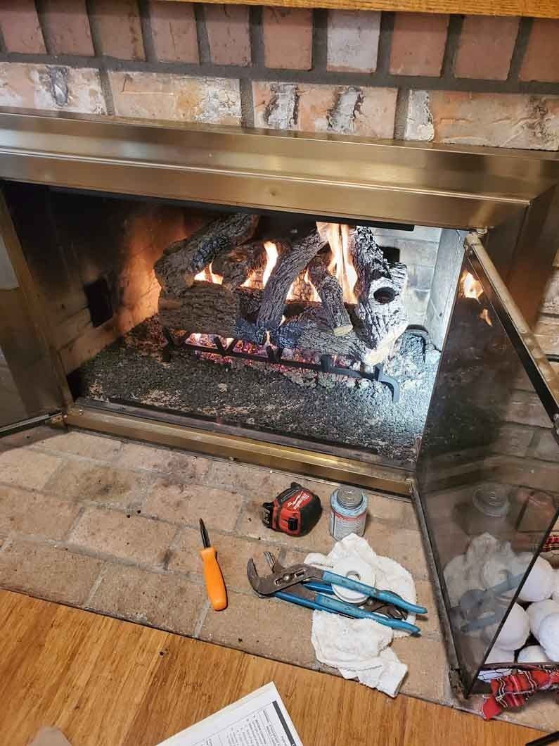 Brick fireplace with a gas log fire burning behind a glass screen, tools and cloth on the hearth.