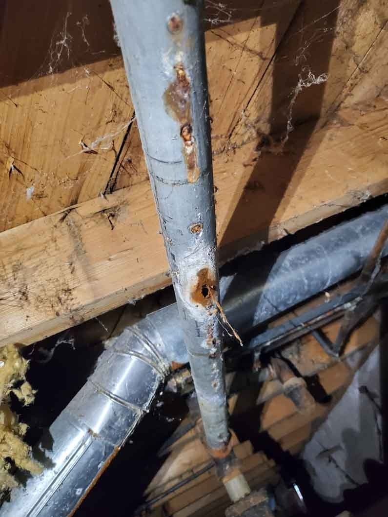Rusty metal pipe and silver HVAC ducts inside a damaged wooden ceiling.