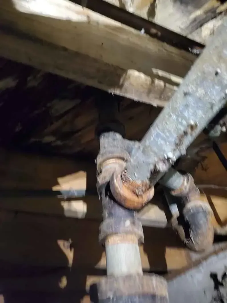 Rusty pipe fittings under a wooden structure, showing corrosion and worn metal connections