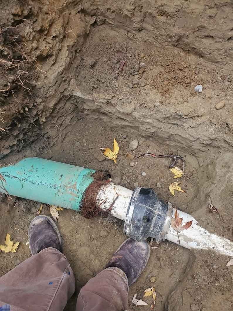 Broken pipe in muddy trench with a person’s boots beside it