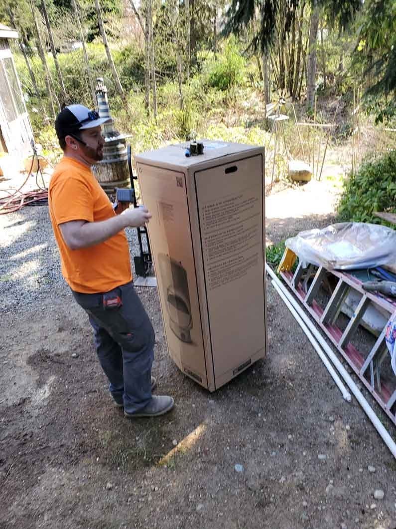 Worker in orange shirt moving a tall cardboard box outdoors beside a ladder and stacked items.