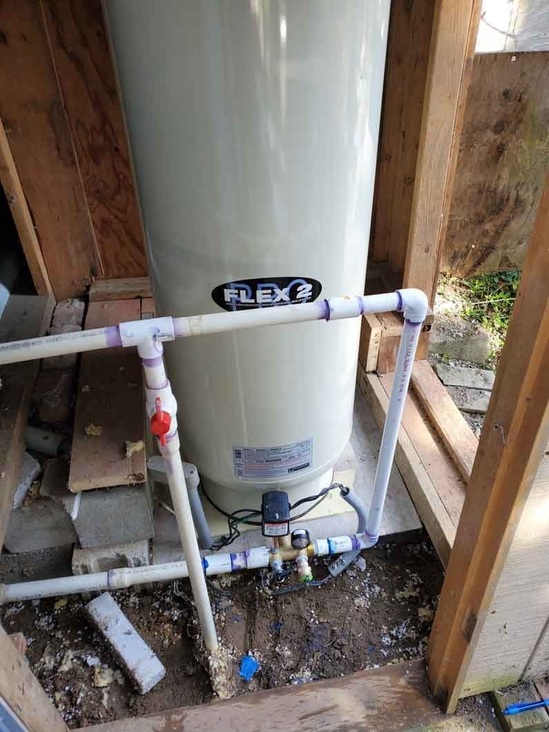 Water heater with white PVC plumbing in a small utility closet, surrounded by wooden framing and dirt floor.