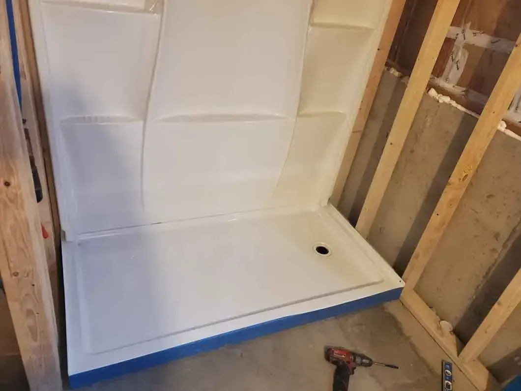 White shower pan installed in an unfinished bathroom, with exposed wall framing and a drill on the floor.